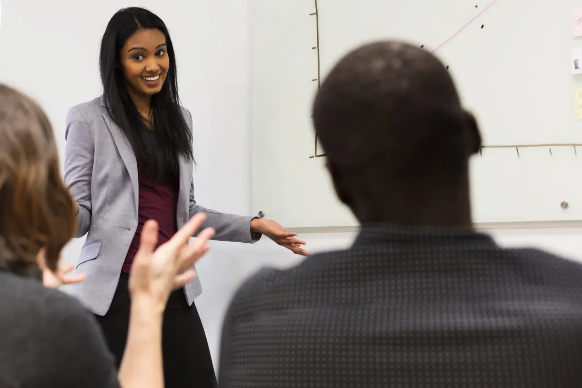 A smiling presenter shows a chart on a whiteboard while a participant in the front row raises a hand